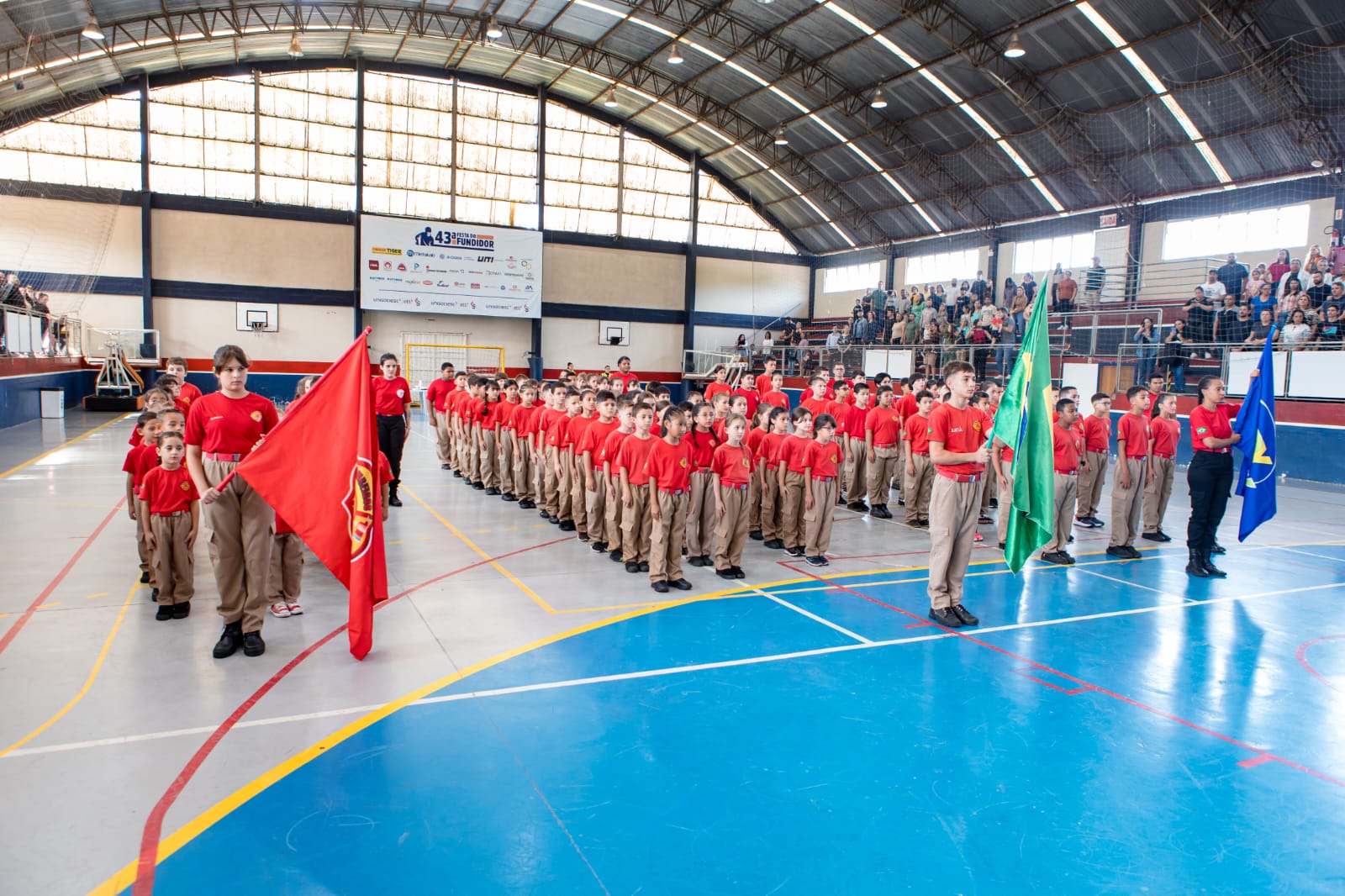 Turmas do Bombeiro Xerife Mirim em formação no ginásio, com as bandeiras do Brasil e do projeto